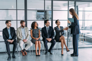 A professional recruiter at a staffing agency shaking hands with a candidate in a modern office waiting area where other diverse job seekers are seated.